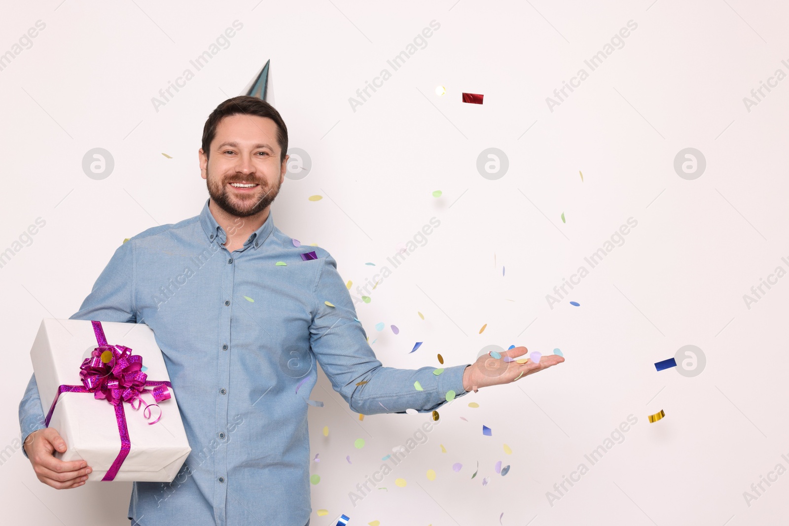 Photo of Happy man with gift box under falling confetti on white background, space for text. Surprise party