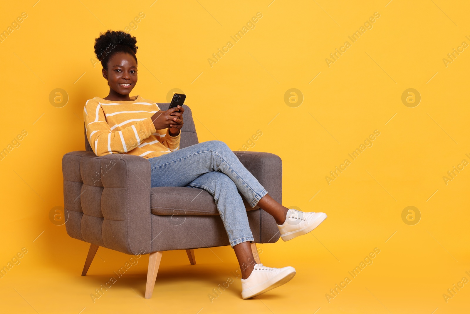 Photo of Smiling woman with smartphone sitting in armchair on yellow background. Space for text