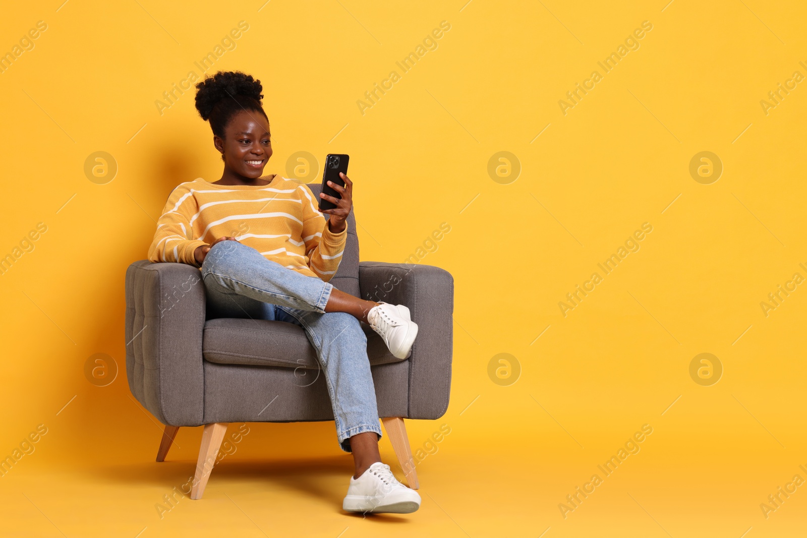 Photo of Smiling woman with smartphone sitting in armchair on yellow background. Space for text