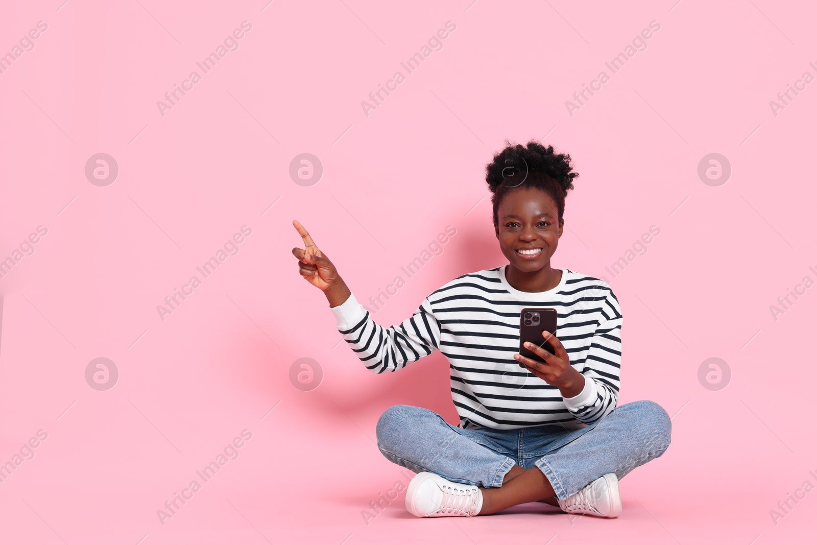 Photo of Cheerful woman with smartphone pointing at something on pink background. Space for text