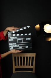 Photo of Woman with clapperboard near spotlight on black background, closeup