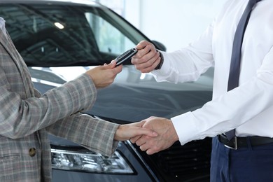 Photo of Salesman giving key to client near new car in salon, closeup