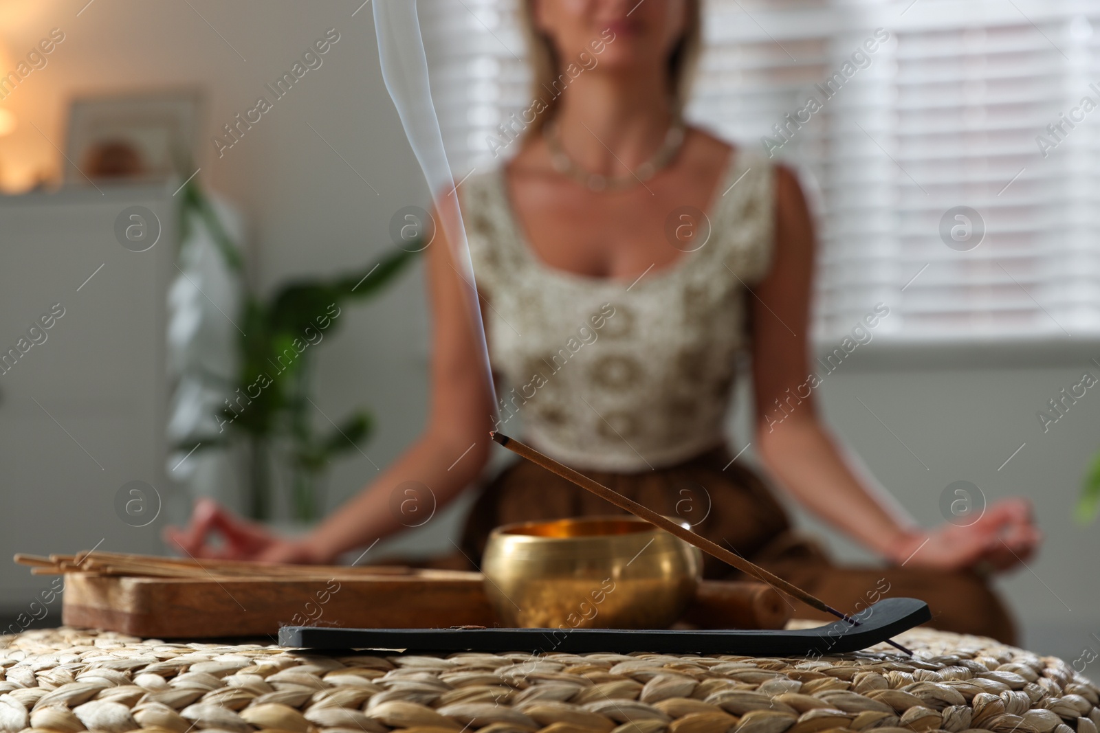 Photo of Young woman practicing yoga indoors, focus on smoldering incense stick and tibetan singing bowl