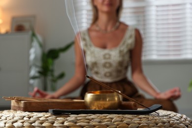 Photo of Young woman practicing yoga indoors, focus on smoldering incense stick and tibetan singing bowl