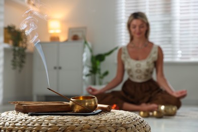 Photo of Young woman practicing yoga indoors, focus on smoldering incense stick and tibetan singing bowl