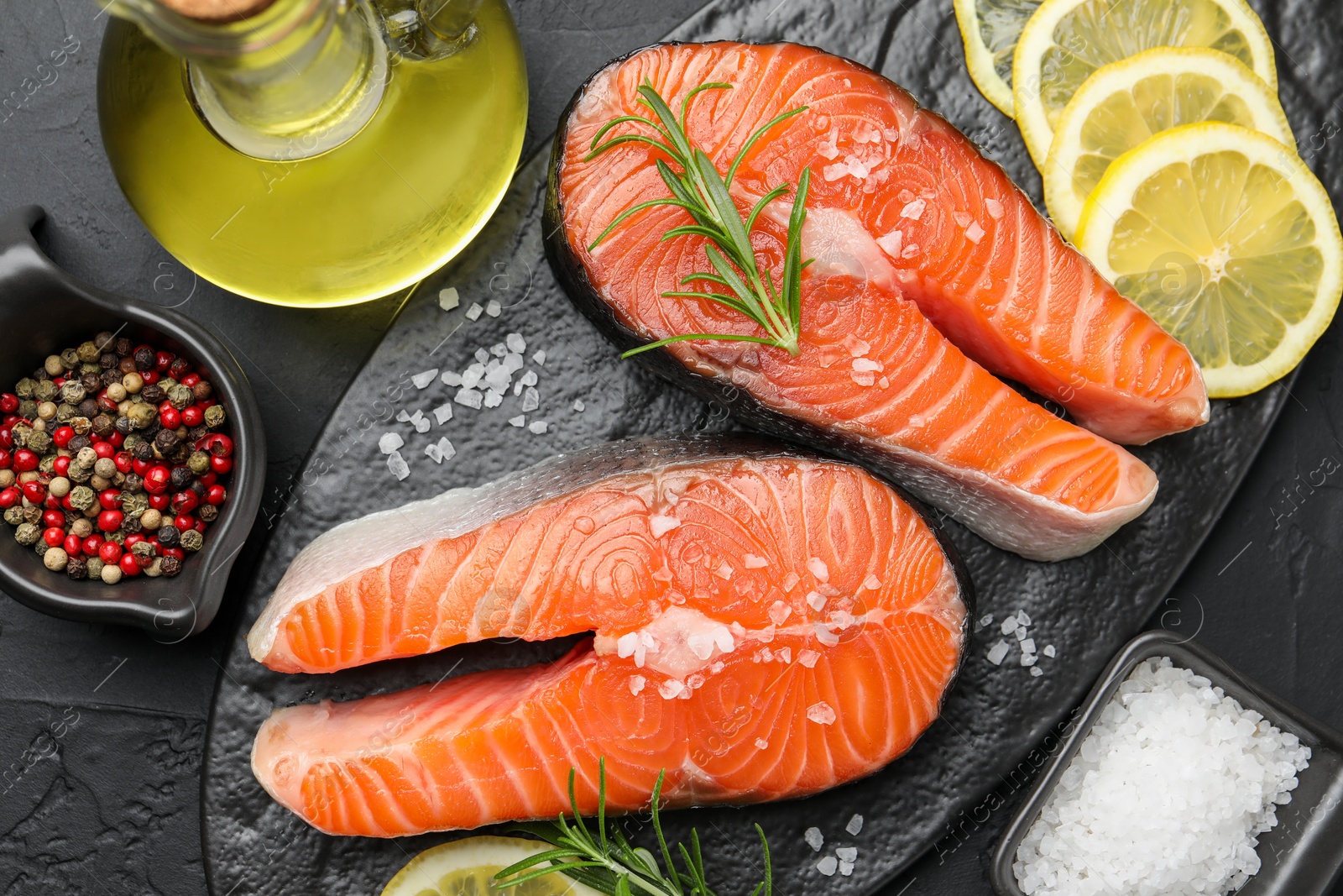 Photo of Fresh raw salmon steaks with salt, lemon, oil, peppercorns and rosemary on dark textured table, flat lay