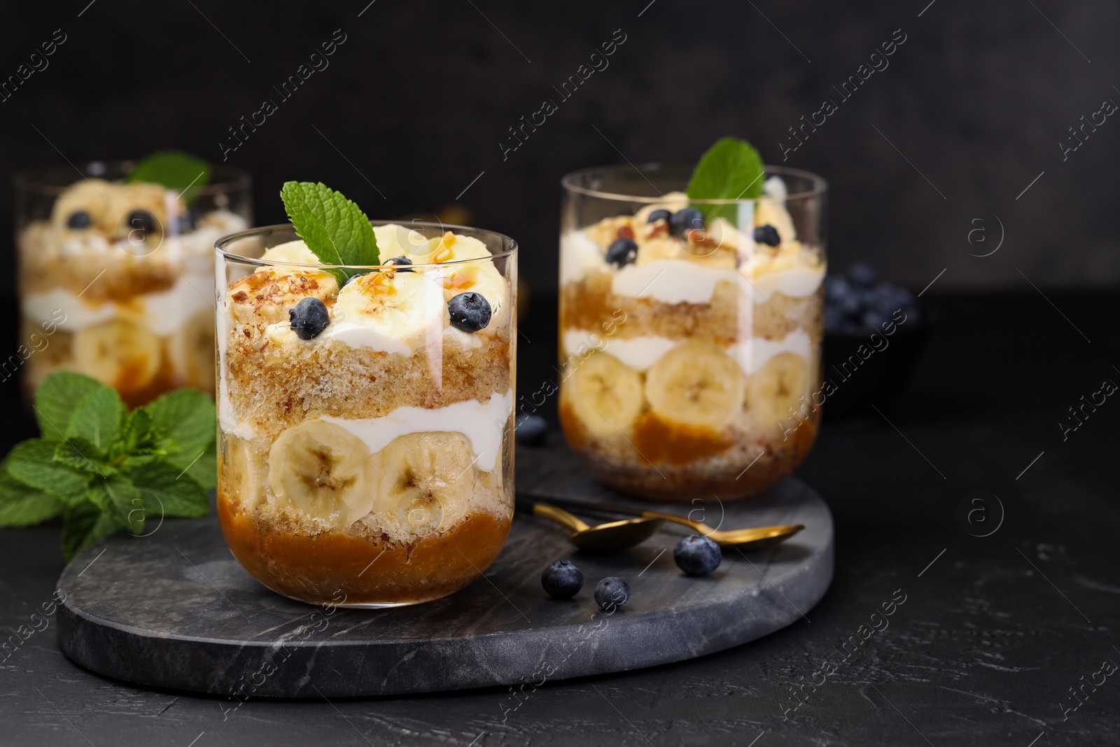 Photo of Tasty trifle dessert. Sponge cake, blueberries, banana and whipped cream in glasses on black table