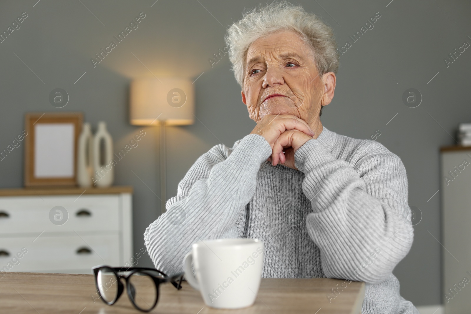 Photo of Sad senior woman feeling lonely at wooden table with cup and glasses indoors