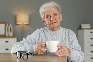 Photo of Sad senior woman feeling lonely at wooden table with cup and glasses indoors