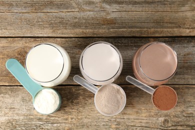 Photo of Different tasty protein cocktails in glasses and powders on wooden table, flat lay