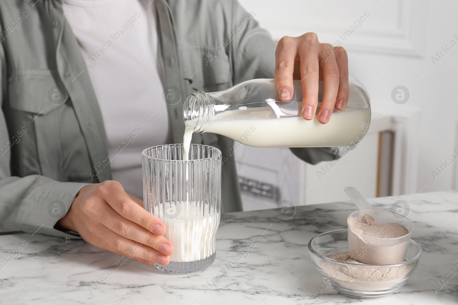 Photo of Making protein cocktail. Woman pouring milk from bottle into glass at white marble table, closeup