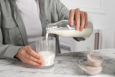 Photo of Making protein cocktail. Woman pouring milk from bottle into glass at white marble table, closeup