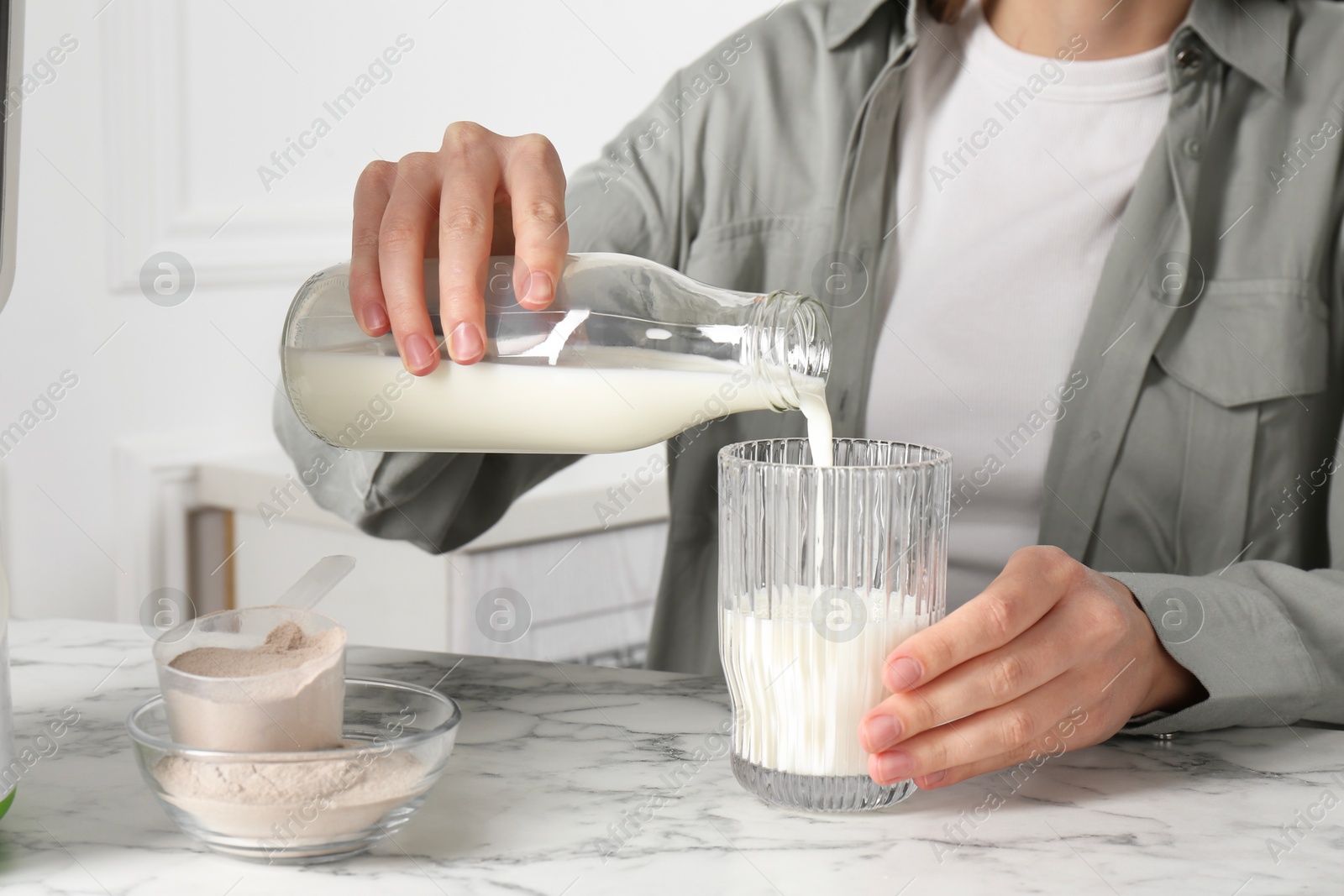 Photo of Making protein cocktail. Woman pouring milk from bottle into glass at white marble table, closeup