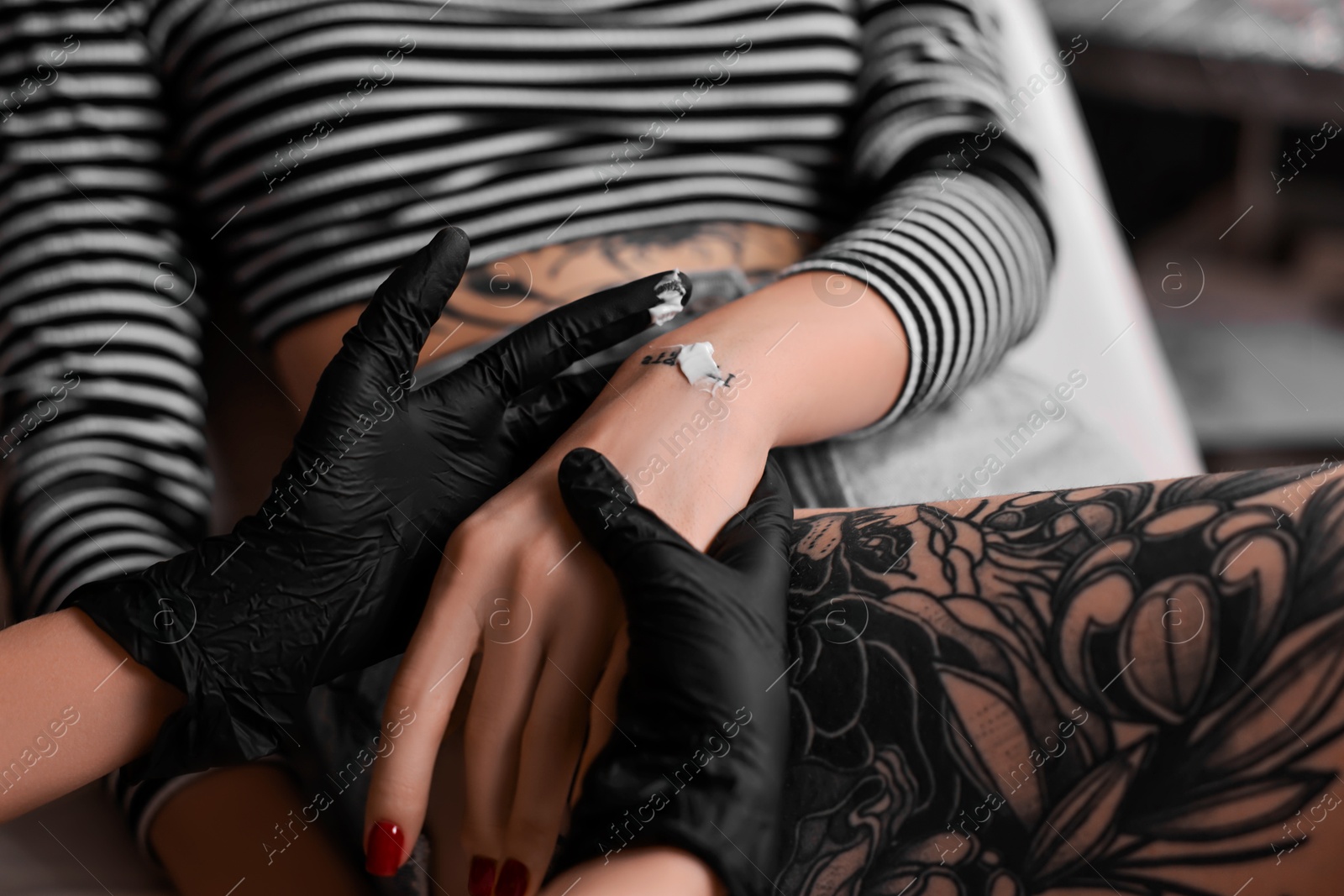 Photo of Professional artist in gloves applying cream onto woman's hand with fresh tattoo in salon, closeup