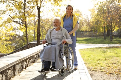 Photo of Caregiver assisting senior man on wheelchair in park. Home health care service