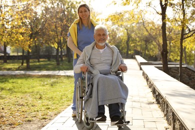 Photo of Caregiver assisting senior man on wheelchair in park. Home health care service