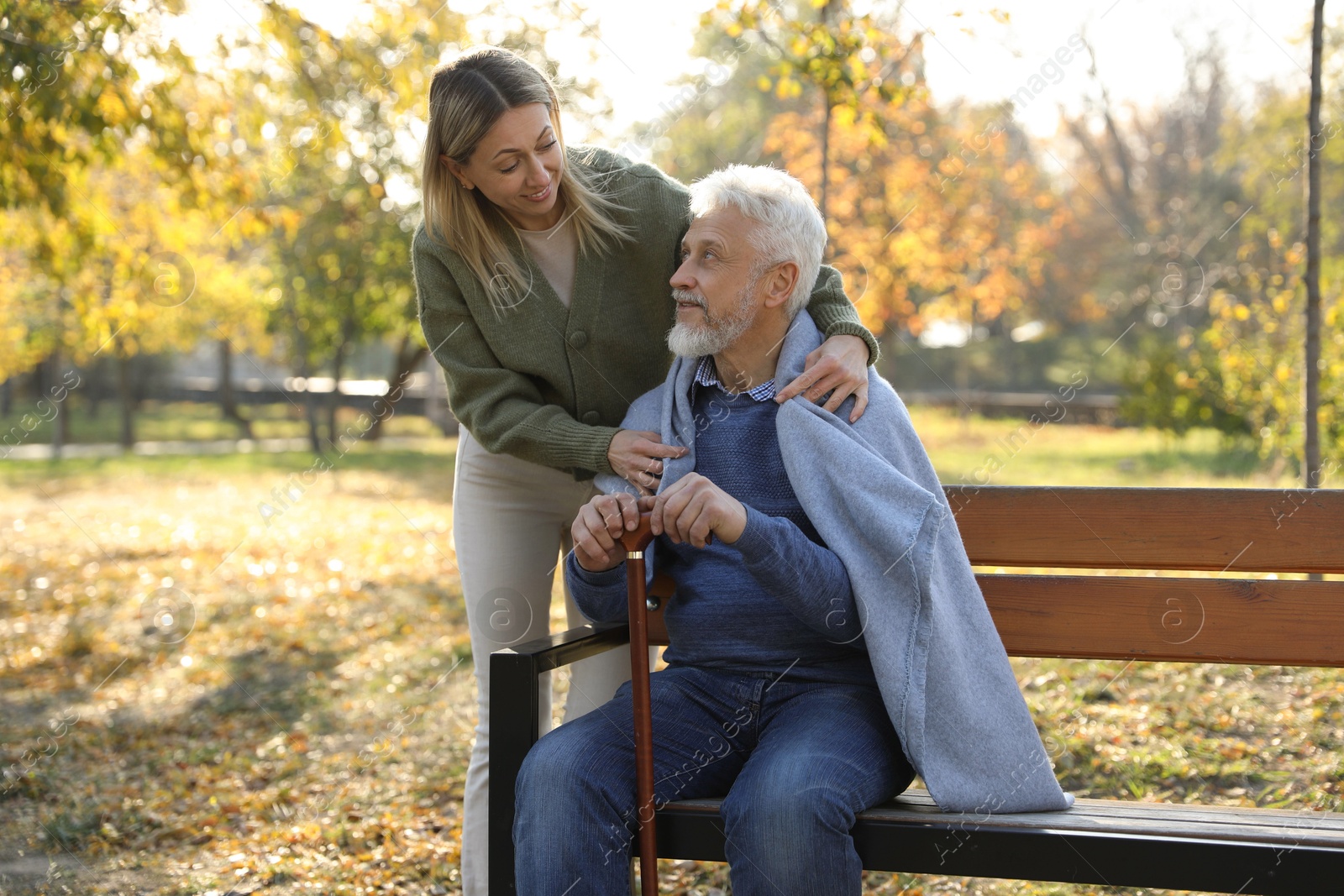 Photo of Caregiver assisting senior man on wooden bench in park. Home health care service