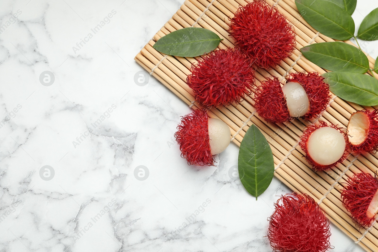 Photo of Delicious ripe rambutans and green leaves on white marble table, flat lay. Space for text