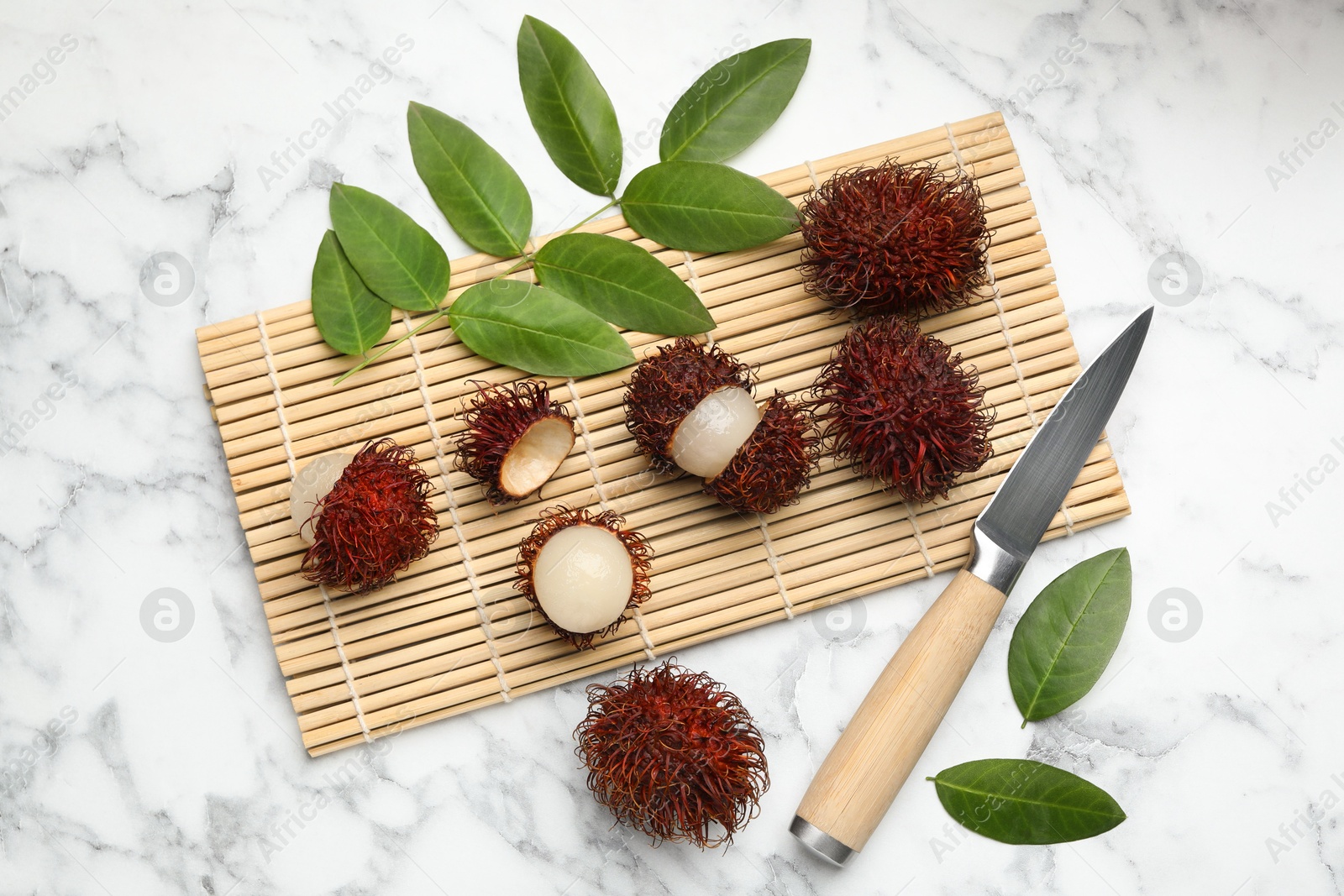 Photo of Delicious ripe rambutans, knife and green leaves on white marble table, flat lay