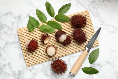 Photo of Delicious ripe rambutans, knife and green leaves on white marble table, flat lay