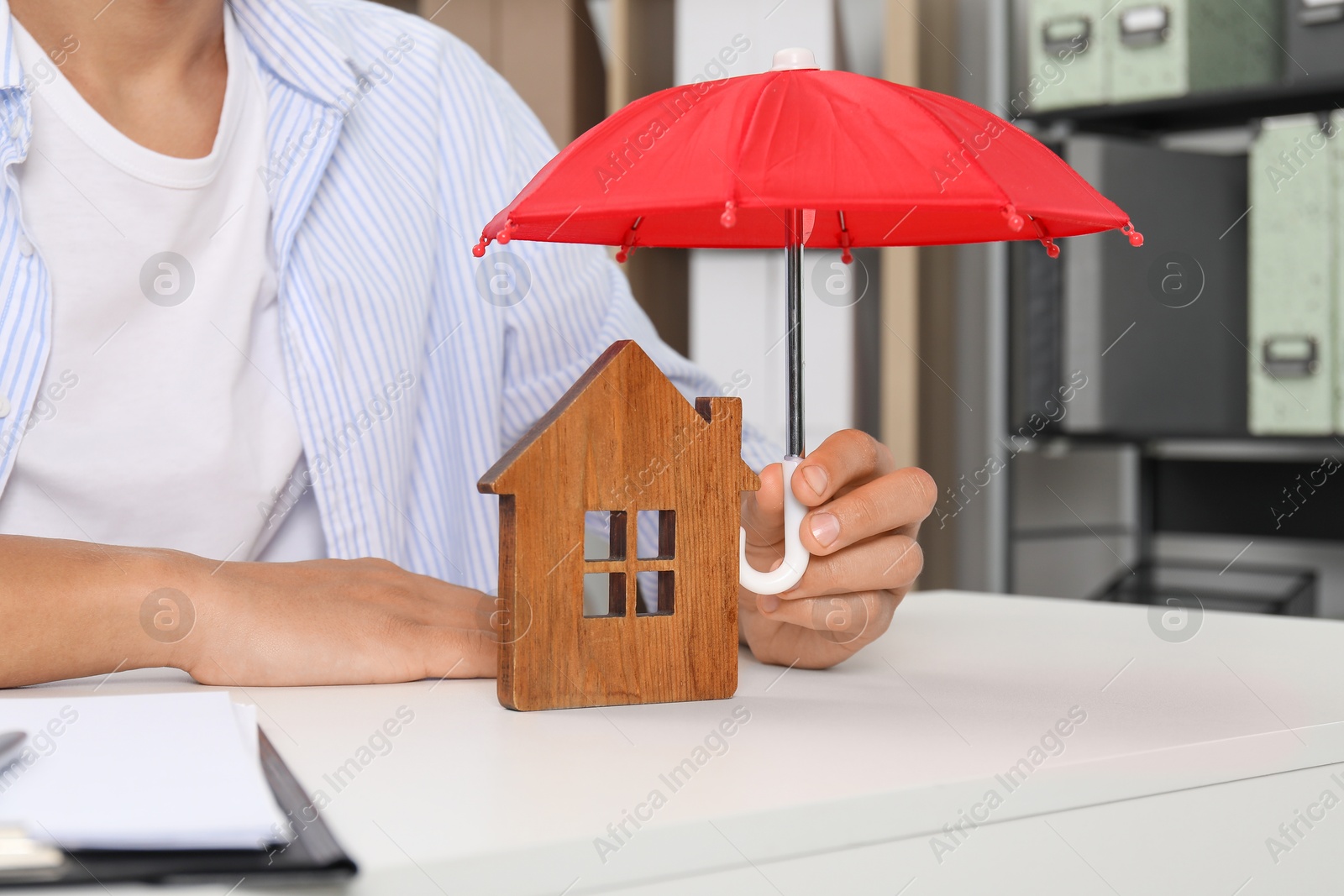 Photo of Real estate insurance. Man holding small umbrella above wooden house figure at light table, closeup