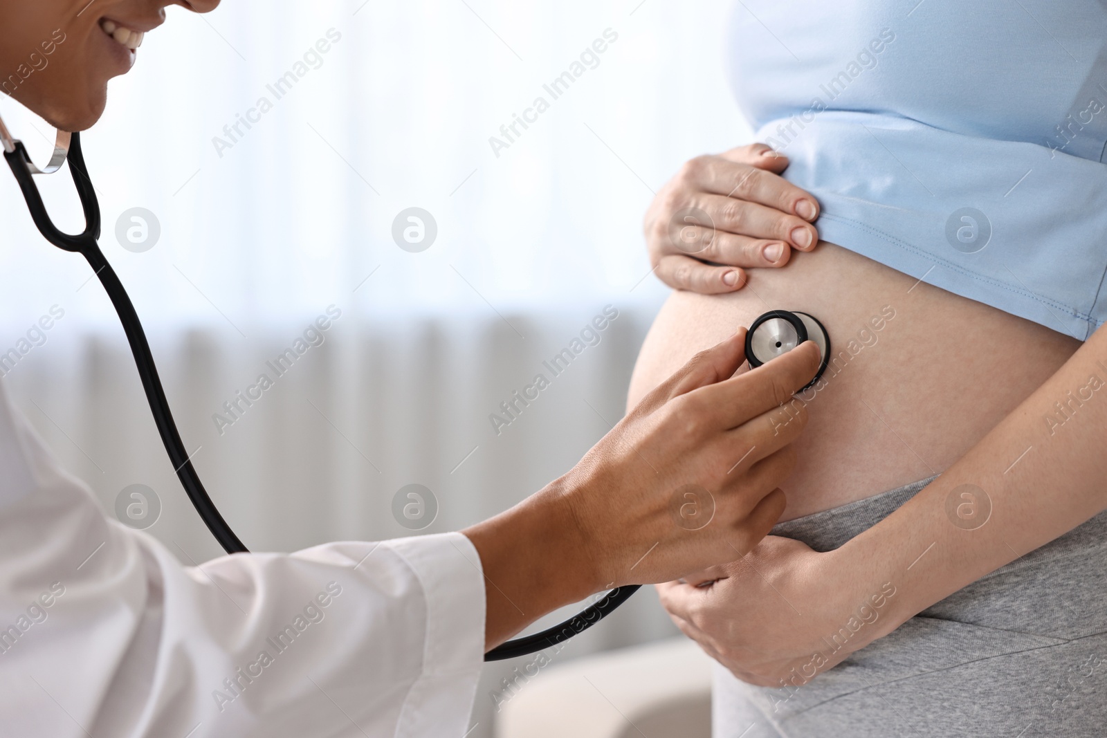 Photo of Pregnancy checkup. Smiling doctor with stethoscope listening baby's heartbeat in patient's tummy indoors, closeup