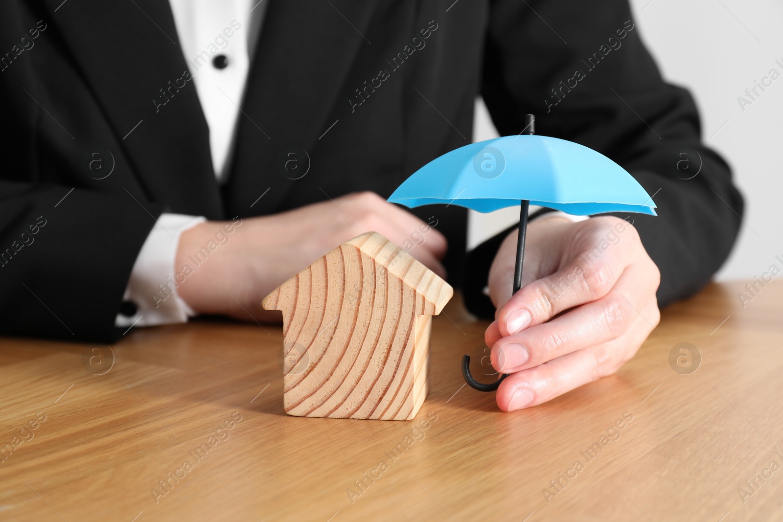 Photo of Property insurance. Real estate agent holding small umbrella above wooden house figure at table indoors, closeup