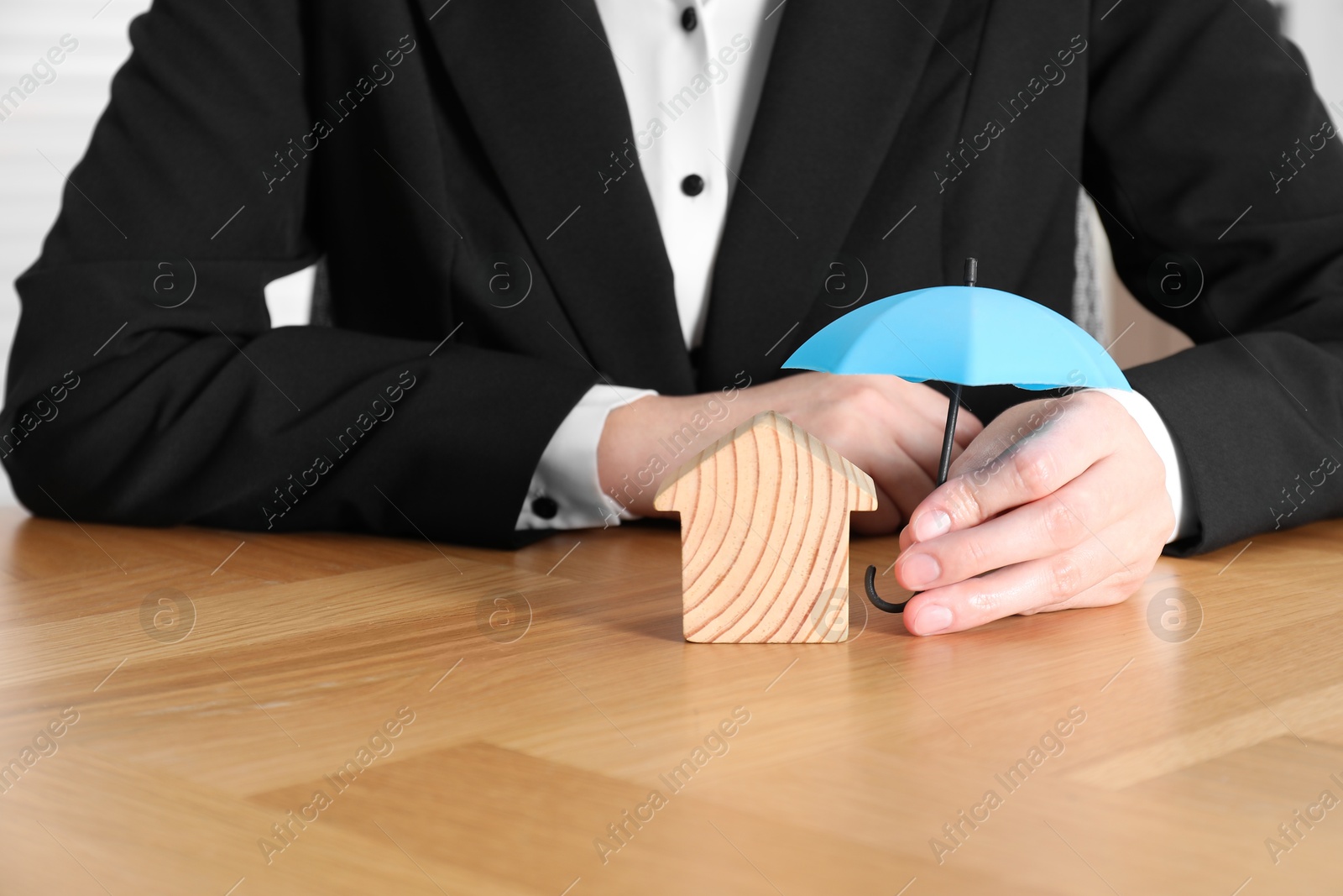 Photo of Property insurance. Real estate agent holding small umbrella above wooden house figure at table indoors, closeup