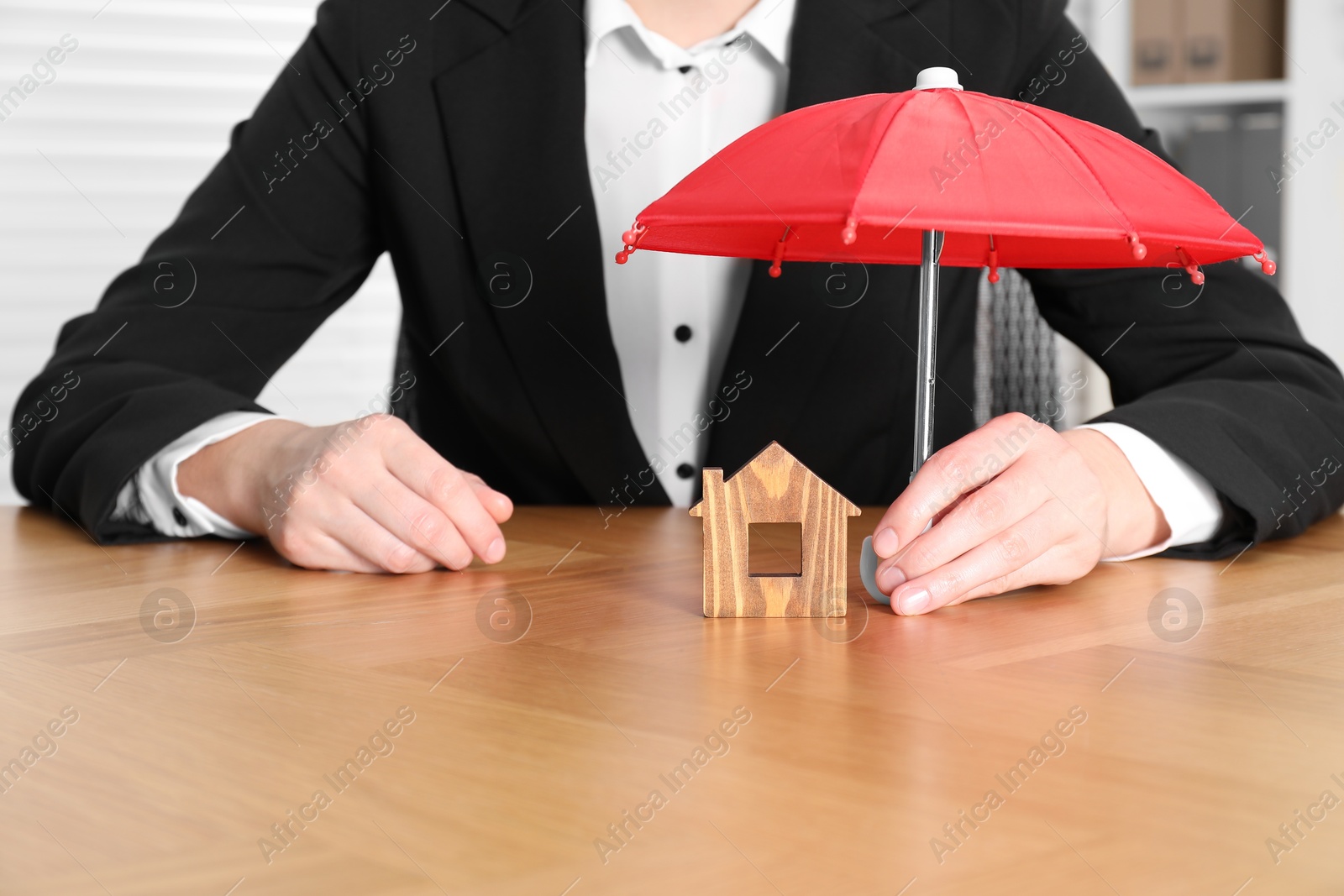 Photo of Property insurance. Real estate agent holding small umbrella above wooden house figure at table indoors, closeup