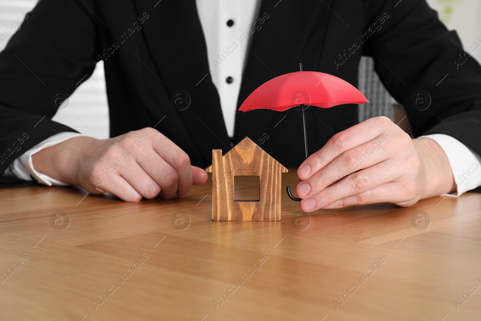 Photo of Property insurance. Real estate agent holding small umbrella above wooden house figure at table indoors, closeup