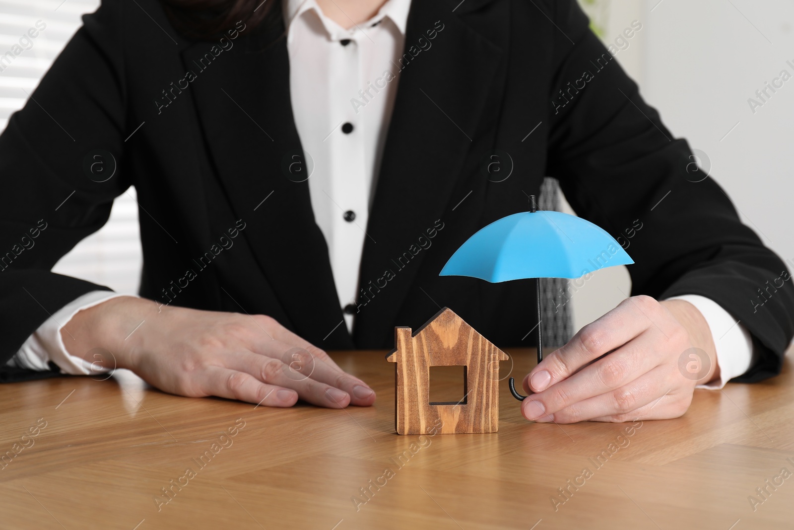 Photo of Property insurance. Real estate agent holding small umbrella above wooden house figure at table indoors, closeup