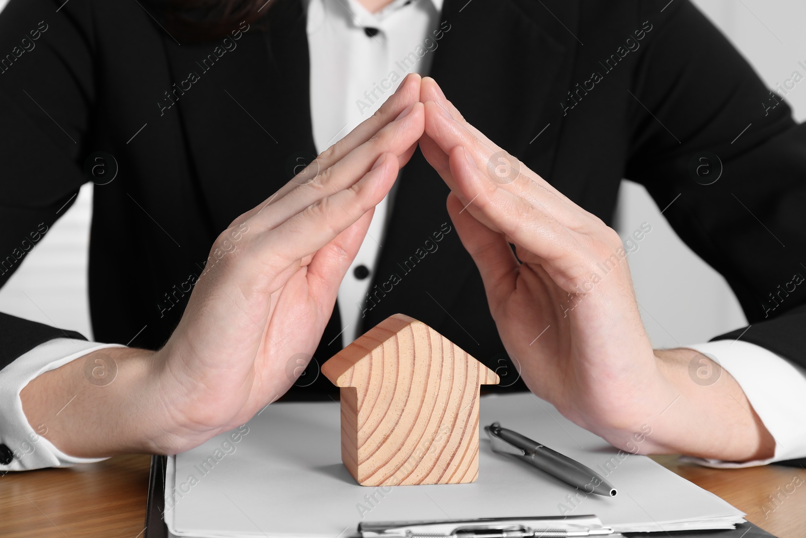 Photo of Property insurance. Real estate agent protecting wooden house figure at table indoors, closeup