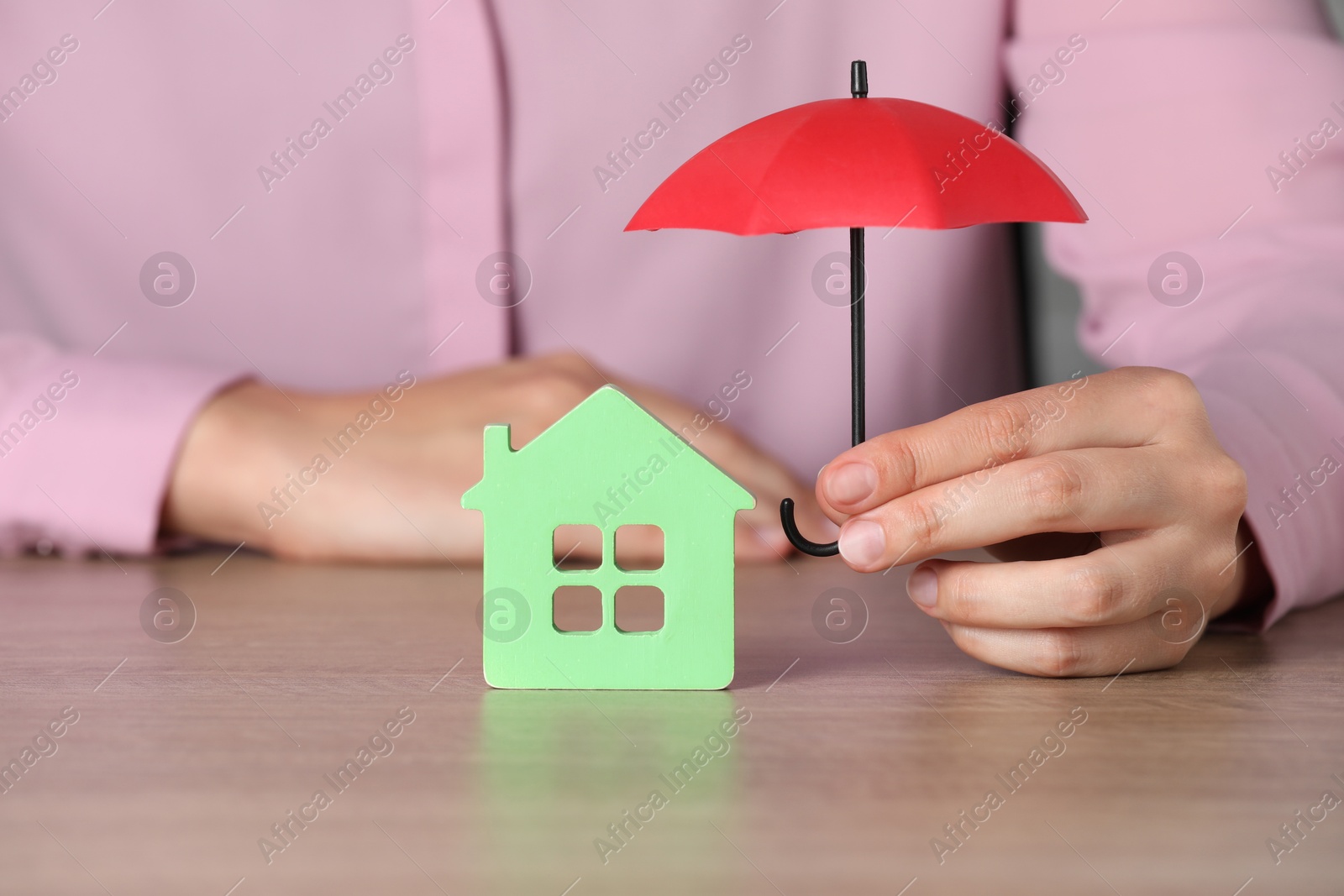 Photo of Property insurance. Real estate agent holding small umbrella above house figure at wooden table indoors, closeup