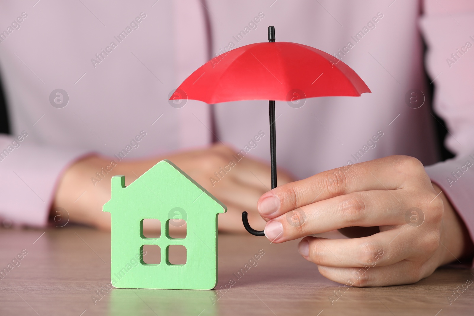 Photo of Property insurance. Real estate agent holding small umbrella above house figure at wooden table indoors, closeup