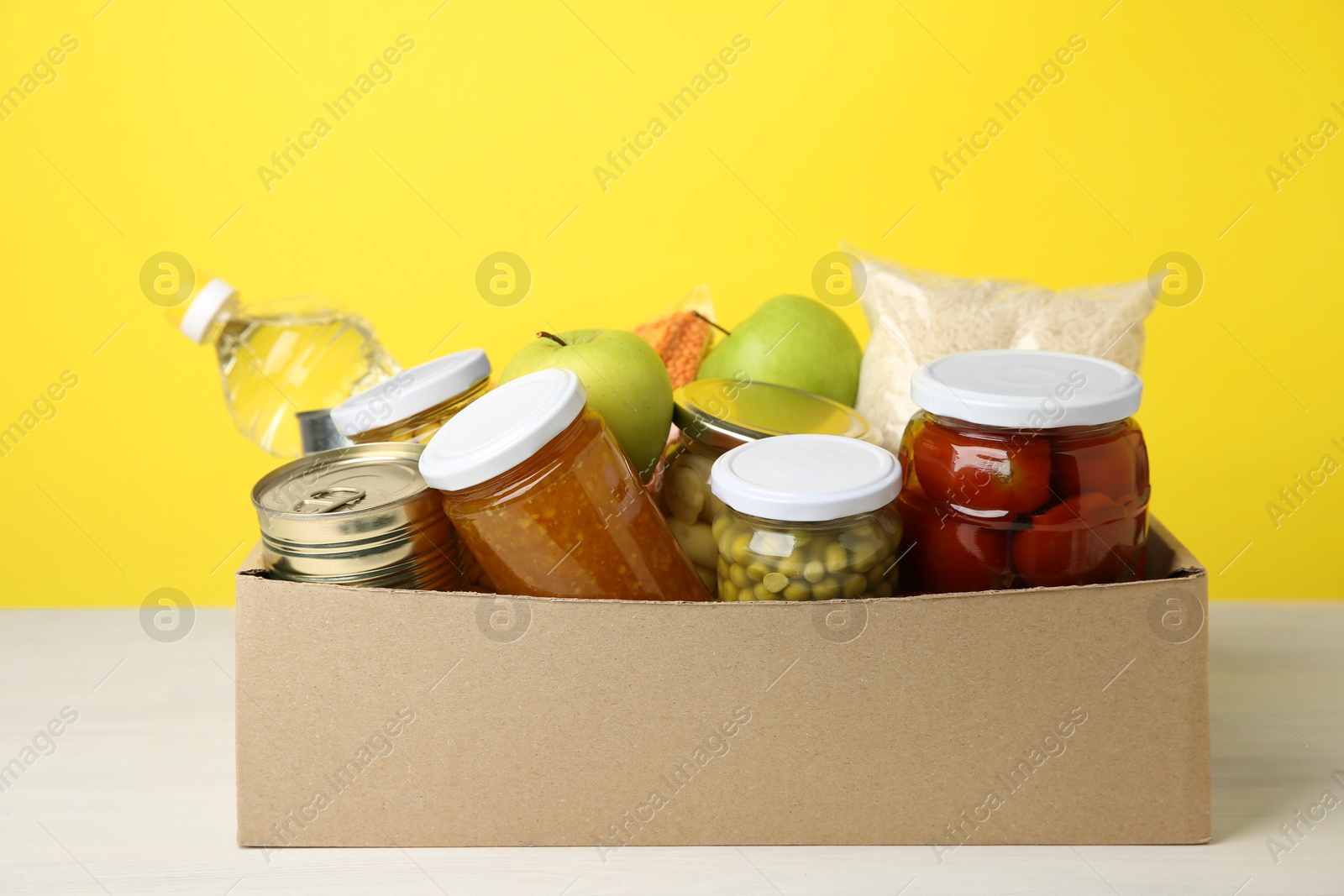 Photo of Different food products for donation in box on table against yellow background