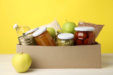 Photo of Different food products for donation in box on table against yellow background