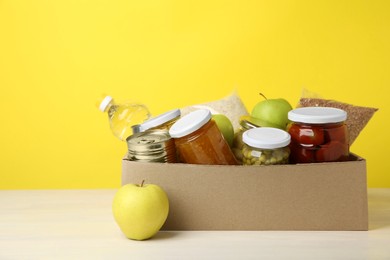 Photo of Different food products for donation in box on table against yellow background