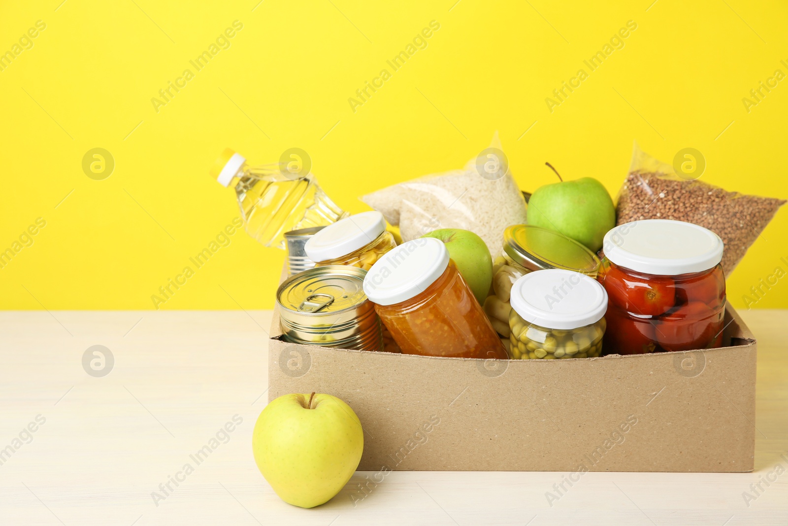 Photo of Different food products for donation in box on table against yellow background, space for text