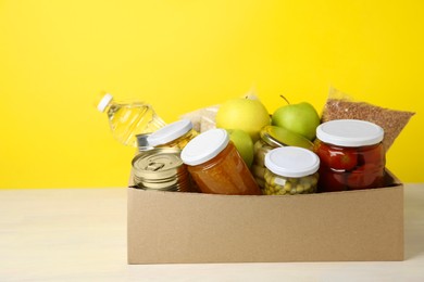 Photo of Different food products for donation in box on table against yellow background