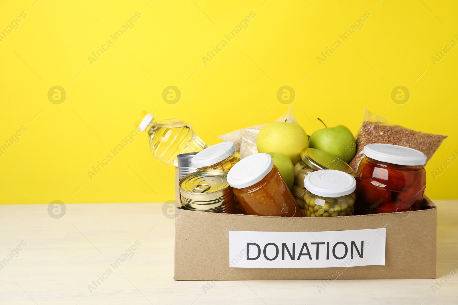 Photo of Different food products for donation in box on table against yellow background, space for text