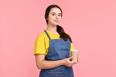 Photo of Girl in apron with paper cup of coffee on pink background. Work for teenagers