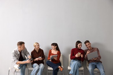 Photo of Group of people using different gadgets on chairs near light grey wall indoors. Modern technology