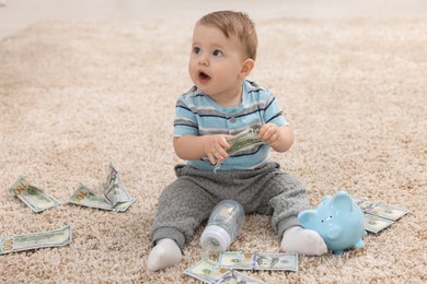 Photo of Little baby with bottle, money and piggybank on floor at home