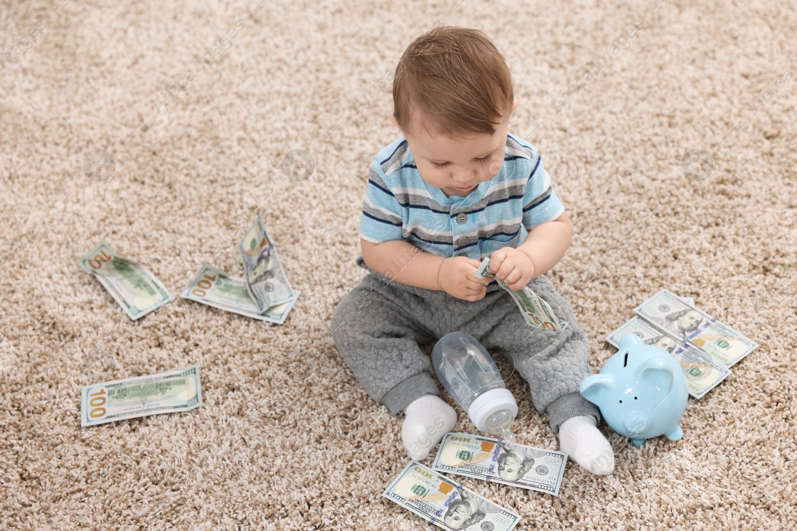 Photo of Little baby with bottle, money and piggybank on floor at home