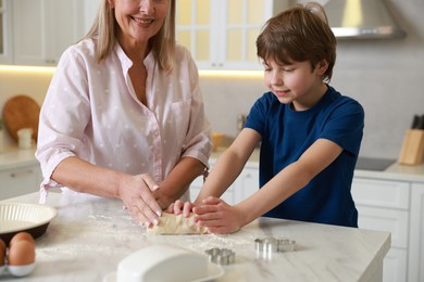 Photo of Grandmother and her grandson kneading dough at white marble table in kitchen