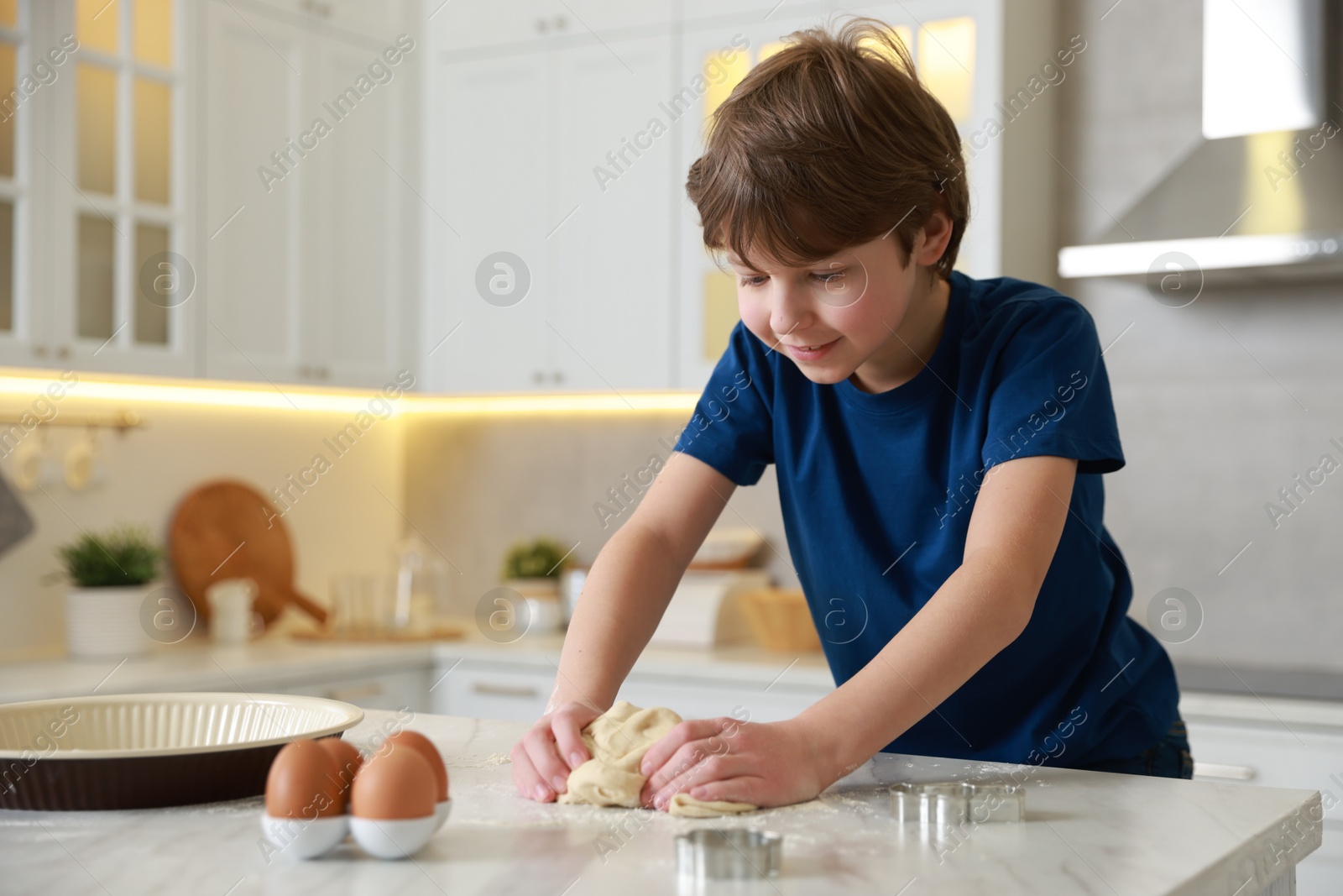 Photo of Cute boy kneading dough at white marble table in kitchen, space for text