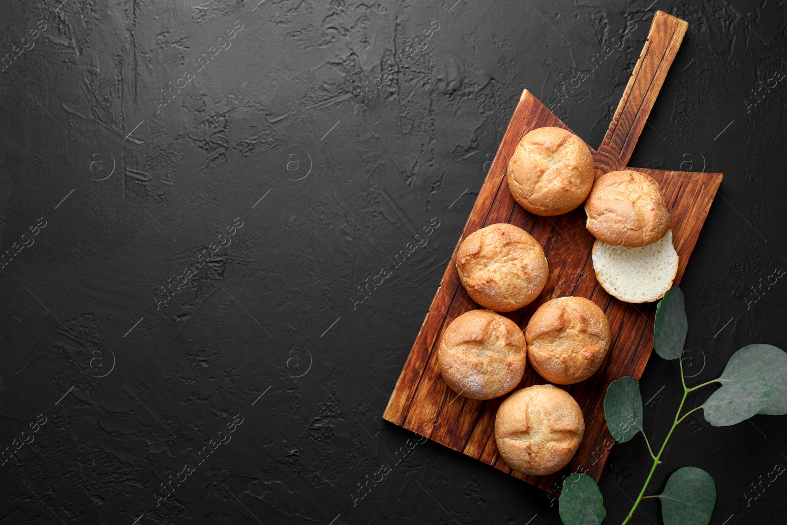 Photo of Homemade tasty buns and eucalyptus branch on black textured table, flat lay. Space for text