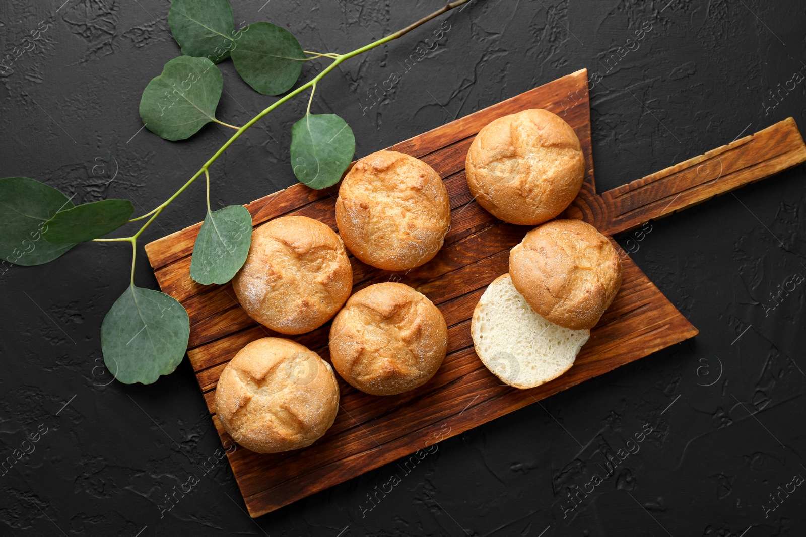 Photo of Homemade tasty buns and eucalyptus branch on black textured table, flat lay