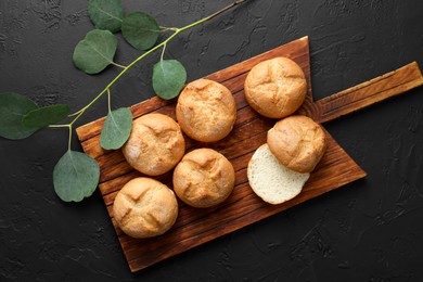 Photo of Homemade tasty buns and eucalyptus branch on black textured table, flat lay