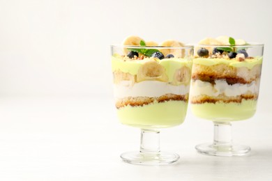 Photo of Tasty trifle dessert. Banana, blueberries, sponge cake and whipped cream in glasses on white wooden table, space for text